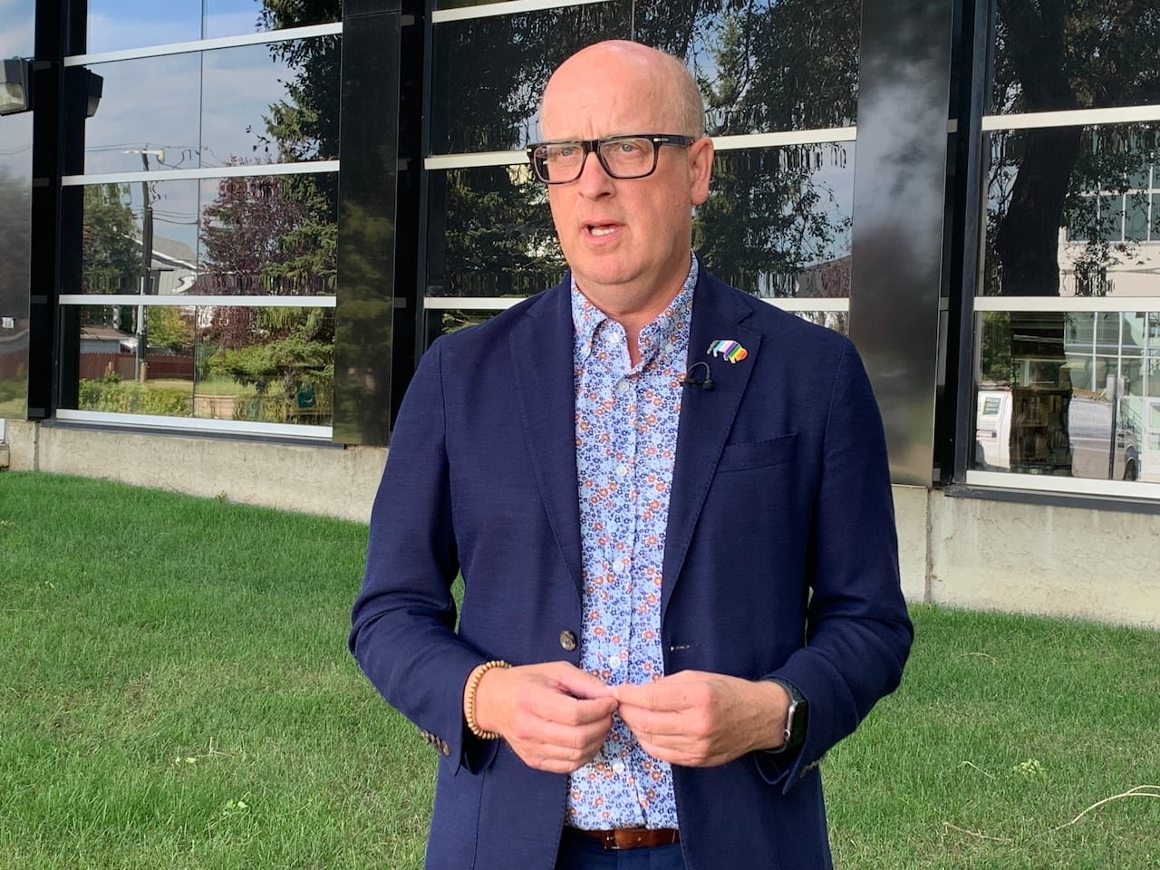 default-416 A man in a navy blazer and flowered dress shirt stands in front of a glass building on a green lawn.